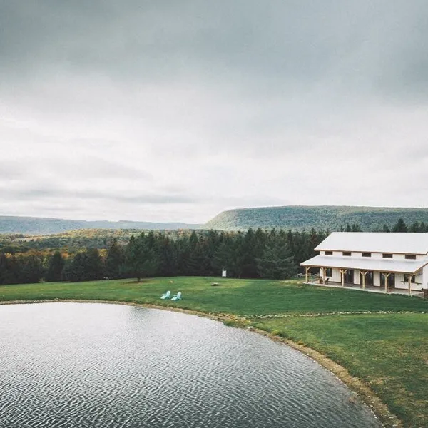 Aerial view of a white barn wedding venue situated on a grassy lawn next to a pond, with a backdrop of forested hills.