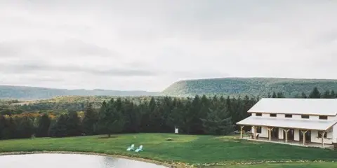 Aerial view of a white barn wedding venue situated on a grassy lawn next to a pond, with a backdrop of forested hills.