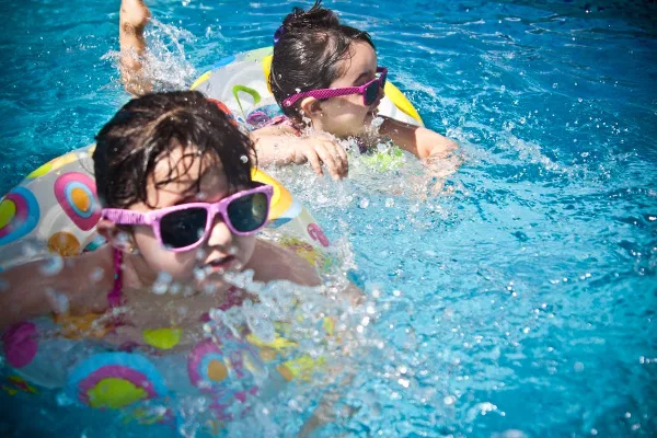 Two children wearing sunglasses and swim rings playing in a blue swimming pool.