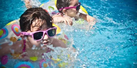 Two children wearing sunglasses and swim rings playing in a blue swimming pool.