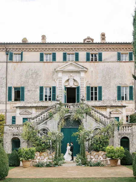 A bride and groom standing at an outdoor wedding ceremony in front of a historic stone building with green shutters and floral arrangements.