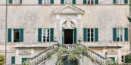 A bride and groom standing at an outdoor wedding ceremony in front of a historic stone building with green shutters and floral arrangements.