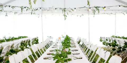 A long white reception table decorated with greenery and string lights under a white tent.