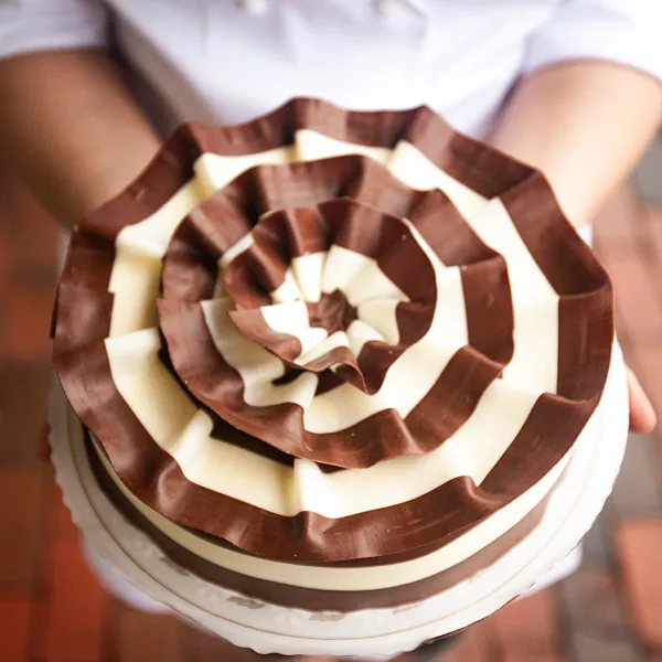 A decorated cake with a spiral pattern of dark and white chocolate ribbons, held by a person.