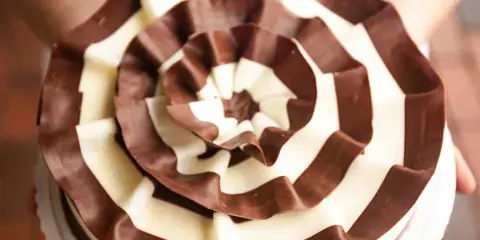 A decorated cake with a spiral pattern of dark and white chocolate ribbons, held by a person.