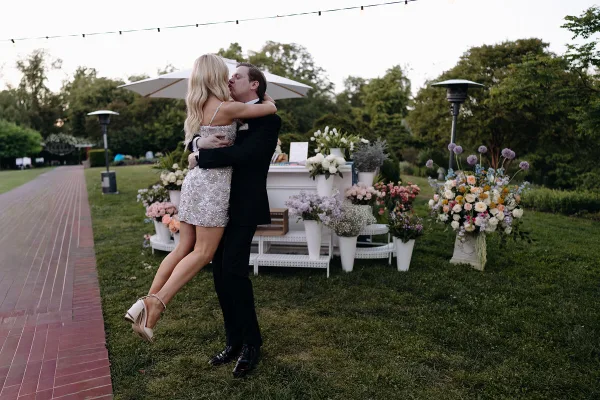 A bride in a sequined dress and a groom in a black suit embrace on a lawn near a floral display.