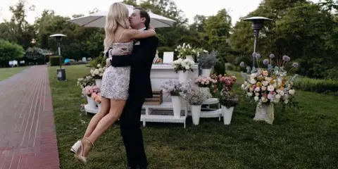 A bride in a sequined dress and a groom in a black suit embrace on a lawn near a floral display.
