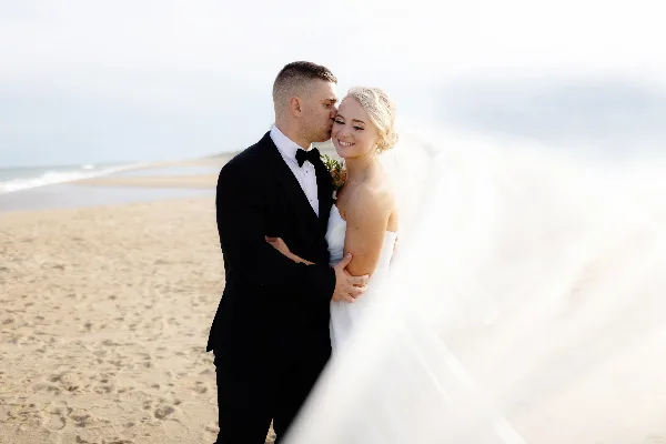 A bride and groom embracing on a sandy beach with the ocean in the background.