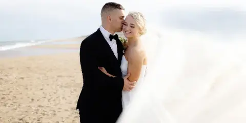 A bride and groom embracing on a sandy beach with the ocean in the background.