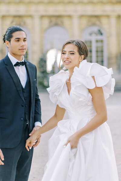 A bride in a white ruffled gown and a groom in a black tuxedo walking together outdoors.