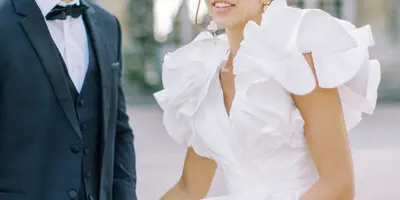 A bride in a white ruffled gown and a groom in a black tuxedo walking together outdoors.