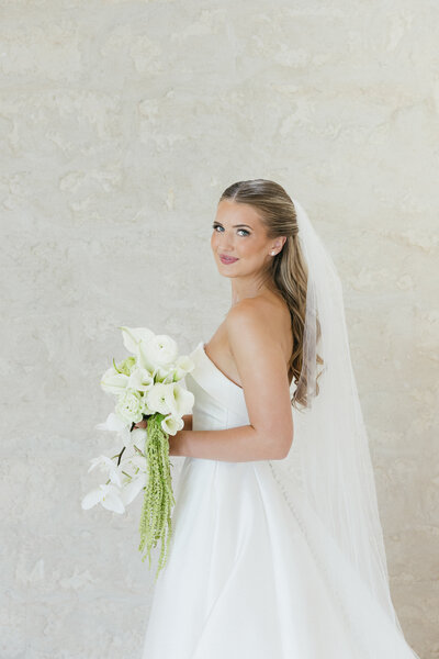 A bride in a white gown and veil holding a bouquet, showcasing a soft-glam hair and makeup style.