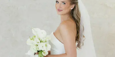 A bride in a white gown and veil holding a bouquet, showcasing a soft-glam hair and makeup style.