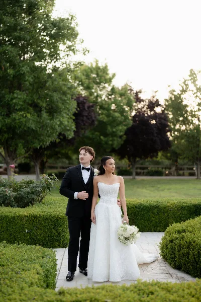 A bride and groom standing together in a garden setting, with the groom in a black tuxedo and the bride in a white wedding dress holding a bouquet.