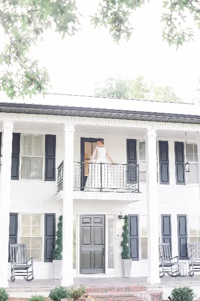 A bride in a white dress standing on the balcony of a white two-story farmhouse venue with dark shutters and rocking chairs on the porch.