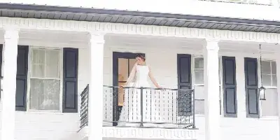 A bride in a white dress standing on the balcony of a white two-story farmhouse venue with dark shutters and rocking chairs on the porch.