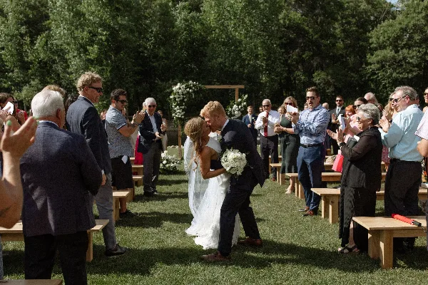 A bride and groom share a kiss during an outdoor wedding ceremony on a lawn, with guests clapping and a floral arch in the background.