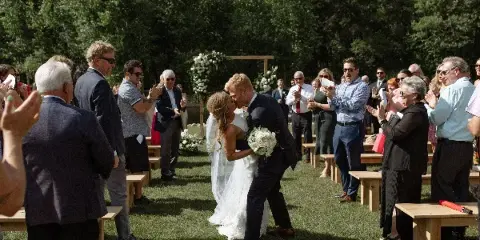 A bride and groom share a kiss during an outdoor wedding ceremony on a lawn, with guests clapping and a floral arch in the background.