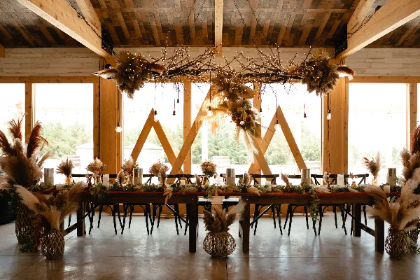 A rustic wedding reception table decorated with floral centerpieces, table runners, and hanging dried floral installations in a wooden venue.