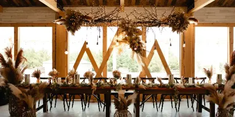 A rustic wedding reception table decorated with floral centerpieces, table runners, and hanging dried floral installations in a wooden venue.
