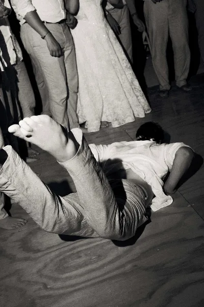 A black-and-white documentary photograph of a person lying on a wooden dance floor with their feet in the air, surrounded by other wedding guests.