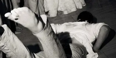 A black-and-white documentary photograph of a person lying on a wooden dance floor with their feet in the air, surrounded by other wedding guests.