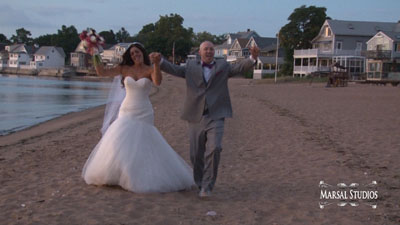 A bride and groom dancing joyfully on a sandy beach near the water, with houses in the background.