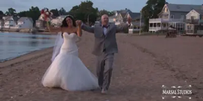A bride and groom dancing joyfully on a sandy beach near the water, with houses in the background.
