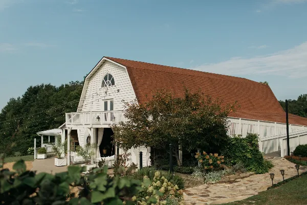 Exterior view of a white barn venue with a red roof, featuring a porch, string lights, and surrounding greenery.