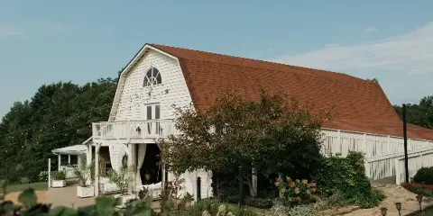 Exterior view of a white barn venue with a red roof, featuring a porch, string lights, and surrounding greenery.