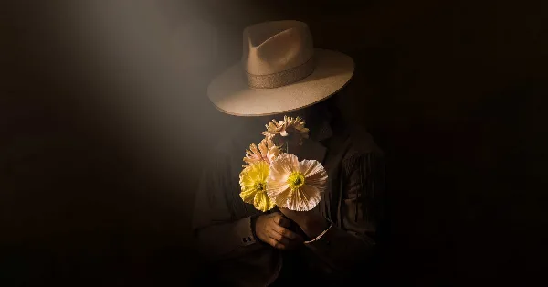 A person in a wide-brimmed hat holding a small bouquet of large, sculptural yellow and white flowers against a dark background.