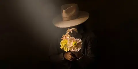 A person in a wide-brimmed hat holding a small bouquet of large, sculptural yellow and white flowers against a dark background.