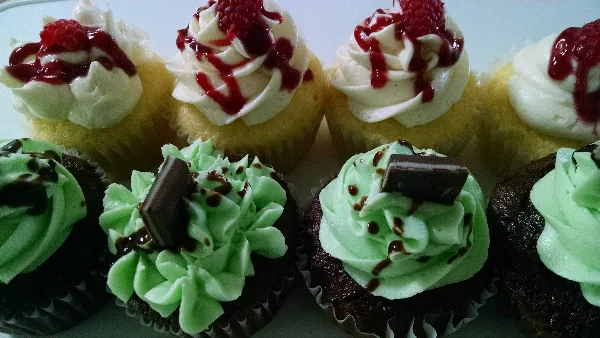 An assortment of gourmet cupcakes featuring white frosting with raspberry sauce and chocolate cupcakes with green frosting and chocolate garnish.