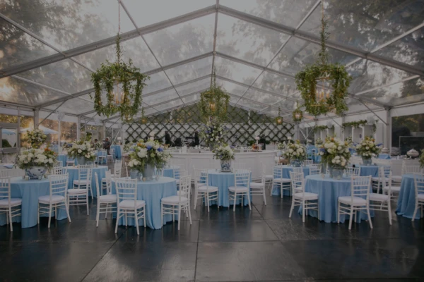 A wedding reception setup inside a clear tent featuring round tables with blue linens, white chairs, and hanging floral installations.