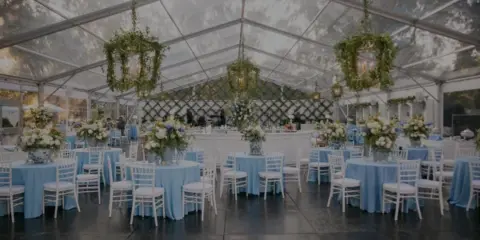 A wedding reception setup inside a clear tent featuring round tables with blue linens, white chairs, and hanging floral installations.