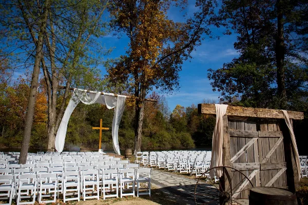 Outdoor wedding ceremony setup with white chairs, a rustic wooden arch, and a cross in a wooded area.