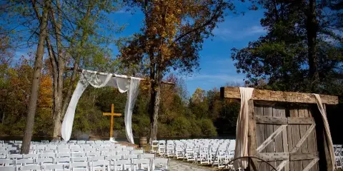 Outdoor wedding ceremony setup with white chairs, a rustic wooden arch, and a cross in a wooded area.