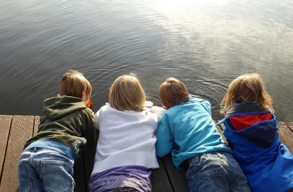 Four children sitting on a wooden dock looking out over a calm body of water.