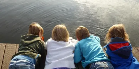 Four children sitting on a wooden dock looking out over a calm body of water.