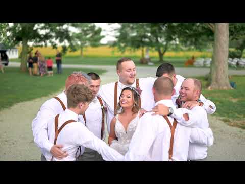 A bride and groom with groomsmen in white shirts and suspenders posing outdoors in a rural setting with trees and a gravel path.