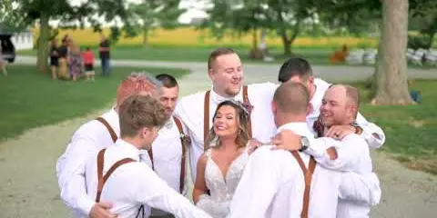 A bride and groom with groomsmen in white shirts and suspenders posing outdoors in a rural setting with trees and a gravel path.