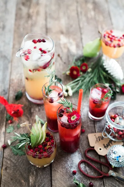 A rustic wooden table setting featuring various colorful infused beverages in glasses and a pitcher, garnished with fresh cranberries, apple slices, and rosemary sprigs.