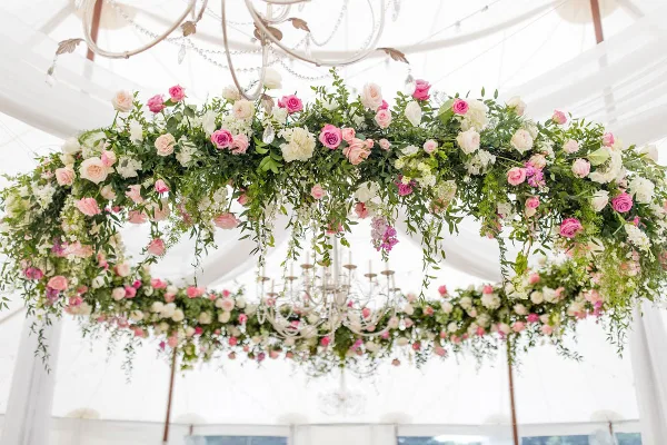 A lush, romantic floral installation with pink and white roses and greenery suspended from a chandelier in a white tent.
