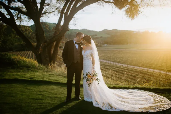 A bride and groom standing close together in a vineyard at sunset, with the bride holding a bouquet and wearing a long veil.