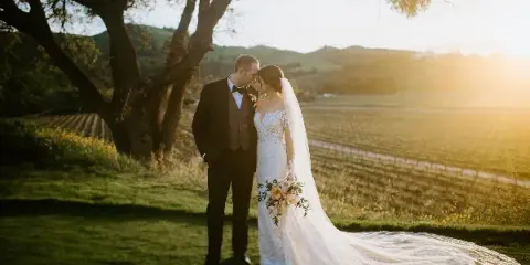 A bride and groom standing close together in a vineyard at sunset, with the bride holding a bouquet and wearing a long veil.