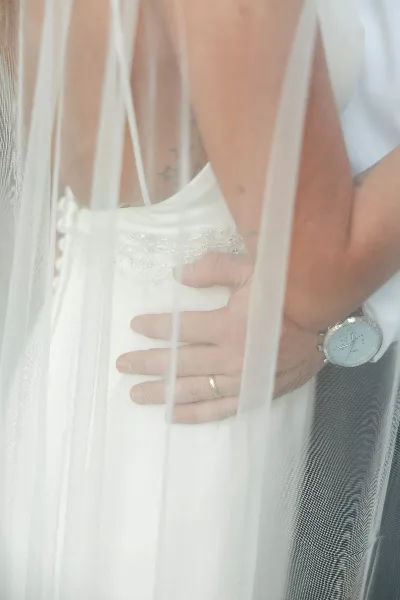 A close-up wedding photograph of a bride and groom embracing, with the groom's hand resting on the bride's waist, partially obscured by the bride's veil.