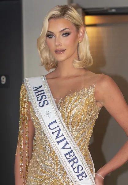 Blonde woman with styled hair and full makeup wearing a gold sequined gown and a Miss Universe sash.