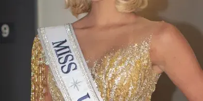 Blonde woman with styled hair and full makeup wearing a gold sequined gown and a Miss Universe sash.