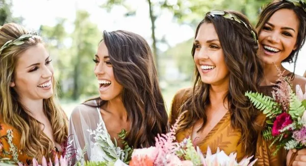 A group of smiling women with styled hair and makeup, holding floral arrangements.