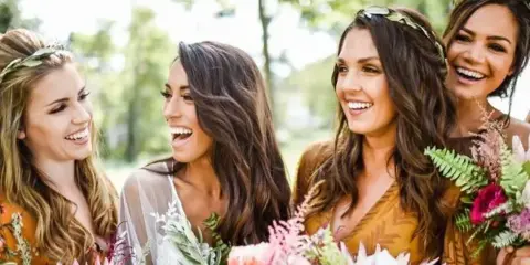 A group of smiling women with styled hair and makeup, holding floral arrangements.